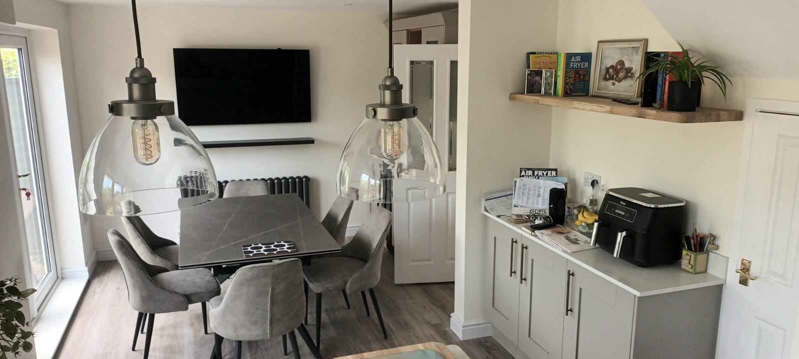 Modern dining room with grey chairs, glass pendant lights, wall-mounted TV, and fitted cabinetry after renovation